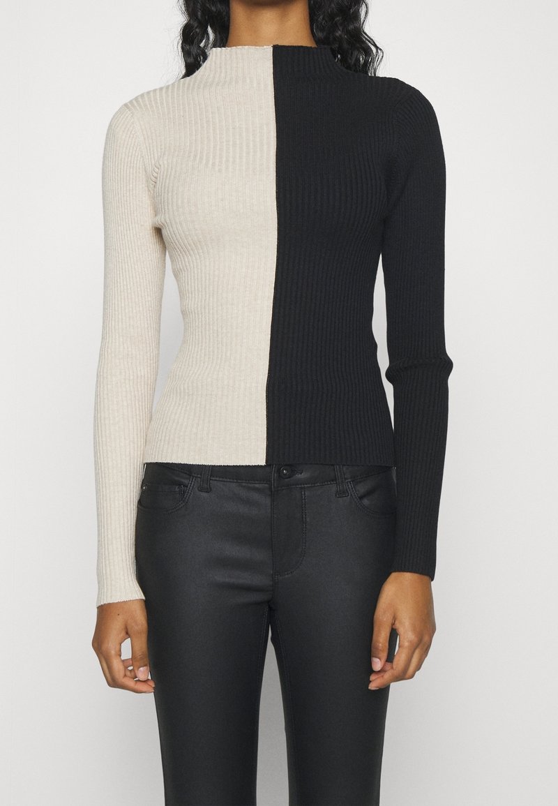 Pull à col roulé côtelé en beige et noir, présentant un design bicolore, des manches longues, et une silhouette ajustée. Porté avec un jean noir.