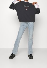 Sudadera de gran tamaño en gris oscuro con el logo de GUESS, combinada con jeans acampanados de color azul claro. La modelo lleva zapatos negros de suela gruesa. Fondo blanco liso.