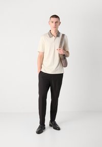 Beige polo skjorte med mønstret krage, kombinert med svarte bukser og svarte loafers. Modellen holder en lysegrå skulderveske.
