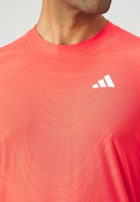 Czerwona koszulka sportowa z teksturowanym wzorem fal i detalami przypominającymi wentylację. Posiada małe białe logo na górnej lewej stronie. Gładka powierzchnia materiału.