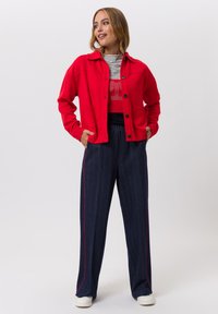 Rote Jacke mit Knöpfen, einem Kragen und zwei Fronttaschen, kombiniert mit navyblauen, gestreiften, weit geschnittenen Hosen mit roten Seitenstreifen. Weiße Sneakers.