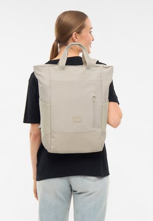 Femme portant un sac à dos rectangulaire beige avec des poignées supérieures, des poches latérales, une poche avant zippée et un petit logo, de dos.