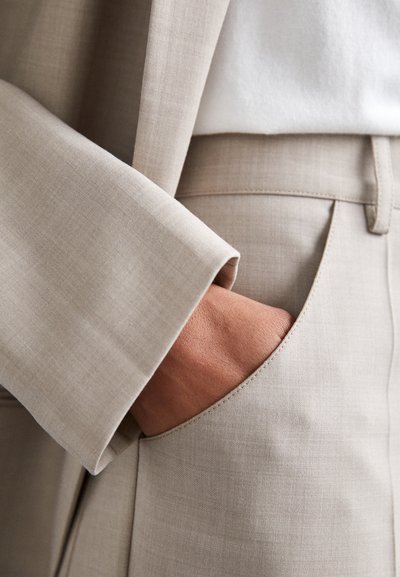 Beige, szabott nadrág sima, textúrált anyagból. Oldalsó zsebbel és gondosan varrt derékrésszel rendelkezik, amelyhez egy világosszürke blazer ujja párosul.