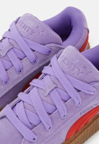 Fialové semišové tenisky s kulatými tkaničkami, gumovými podrážkami a červenými akcenty. Obsahují ražební značení na boku a texturovaný límec.