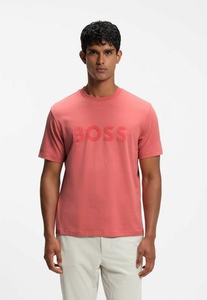 Mann trägt ein korallenrosa T-Shirt mit dem Text "BOSS" und hellbeiger Hose, steht vor einem schlichten weißen Hintergrund.