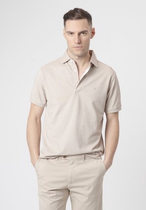 Hombre con cabello corto y oscuro, vestido con camiseta tipo polo beige de manga corta y pantalones beige a juego, de pie con las manos en los bolsillos sobre un fondo blanco.