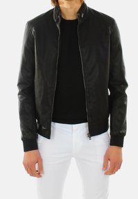 Giubbotto bomber in pelle nera con zip frontale, polsini aderenti e colletto rotondo. Indossato sopra una camicia scura e pantaloni bianchi.