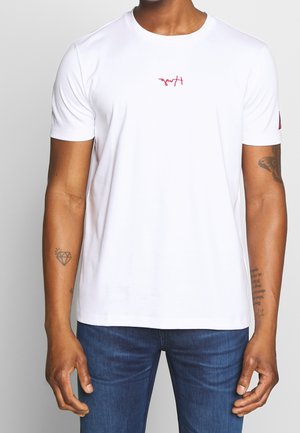 Mann trägt schlichtes weißes kurzärmeliges T-Shirt mit kleinem roten Schriftzug auf der Brust und blauen Jeans, zeigt Tattoos auf beiden Unterarmen.