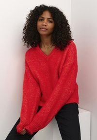 Maglione rosso a maglia con scollo a V e maniche oversize, con una finitura testurizzata. Indossato con pantaloni neri, su uno sfondo chiaro.
