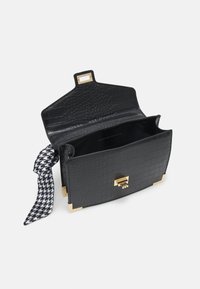 Sac à main en cuir noir texturé avec protège-coins dorés et fermoir, rabat supérieur ouvert, et tissu pied-de-poule noir et blanc noué sur le côté.