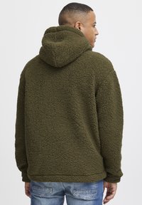 Green fleece hoodie with a textured surface, featuring a hood and a relaxed fit, paired with light blue jeans.