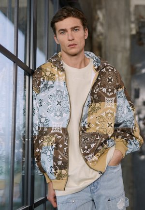 Reißverschluss-Hoodie mit einem mehrfarbigen Paisleymuster in Braun, Blau und Beige; darunter sichtbar ein helles T-Shirt und Jeans-Shorts.