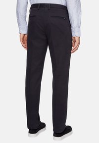 Black, fitted trousers with two rear zip pockets. The texture appears smooth, made from a durable fabric. Worn with a light blue striped shirt.