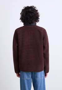 Brown textured fleece jacket with a black collar, featuring a relaxed fit and raglan sleeves. Worn over light blue wide-leg jeans.