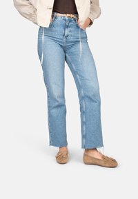 Persoon die lichtblauwe high-waist jeans met rafelige zoom, tan loafers met kwastjes, een beige jas en een zwart shirt draagt, staand met handen in de zakken.