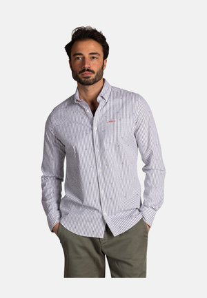 Homme barbu portant une chemise à rayures gris clair et blanc boutonnée et un pantalon vert olive, mains dans les poches, arrière-plan neutre.