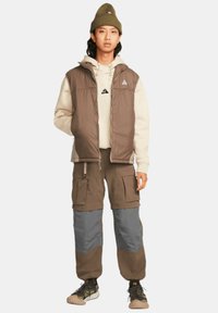 Brun pufferväst över en beige hoodie, i kombination med cargo-byxor med grå paneler. En texturerad mössa och mönstrade sneakers fulländar looken.