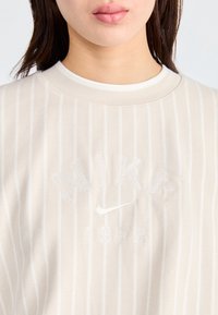 Närbild av en person som bär en beige och vit vertikalt randig Nike-tröja med broderad logotyp och texten "1972" på framsidan.