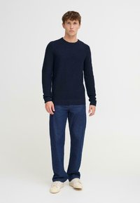 Donkerblauwe gebreide trui met een ronde halslijn en lange mouwen, gedragen met blauwe spijkerbroek en lichtgekleurde sneakers. Eenvoudig ontwerp.