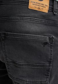 Mustat farkut, joissa on takatasku vaakasuoralla ompeleella ja ruskea nahkainen etiketti, jossa lukee "DENIM DRIVEN SINCE 1989."