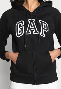 Sweat à capuche zippé noir en tissu doux avec des poignets côtelés, arborant un grand logo "GAP" blanc sur la poitrine et des poches avant.
