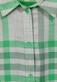 Camicia a scacchi verde e grigia con un tessuto testurizzato, colletto e bottoni verdi. La cucitura visibile aggiunge dettagli sulla parte anteriore.