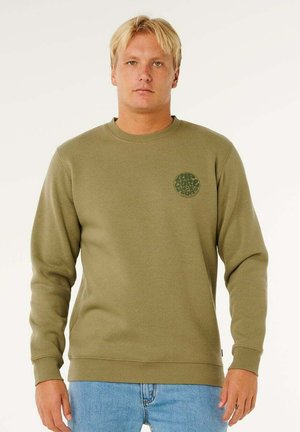 Blondyn w oliwkowozielonej bluzie z długim rękawem z ciemnozielonym okrągłym logo na piersi i jasnoniebieskich dżinsach, stojący na białym tle.