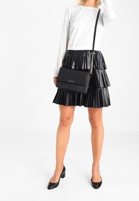 Schwarze Leder-Umhängetasche mit Kettenriemen, rechteckiger Form und minimalistischem Design. Kombiniert mit einem weißen Langarmoberteil und einem plissierten schwarzen Rock.