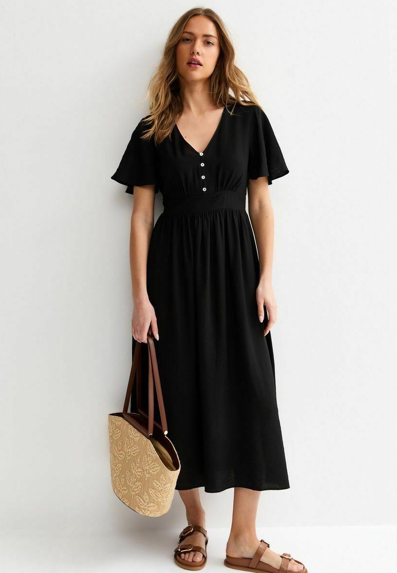 Femme portant une robe midi noire avec des boutons et des manches volantées, tenant un sac tote beige, debout contre un mur blanc.