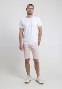 T-shirt blanche à manches courtes avec un petit logo, accompagnée de shorts roses clairs et de baskets blanches avec des semelles beiges, se tenant contre un fond uni.