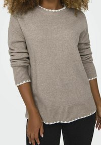 Hellbrauner Strickpullover mit geripptem Saum und gewelltem weißen Besatz um den Halsausschnitt und die Bündchen, aus weichem Stoff gefertigt.