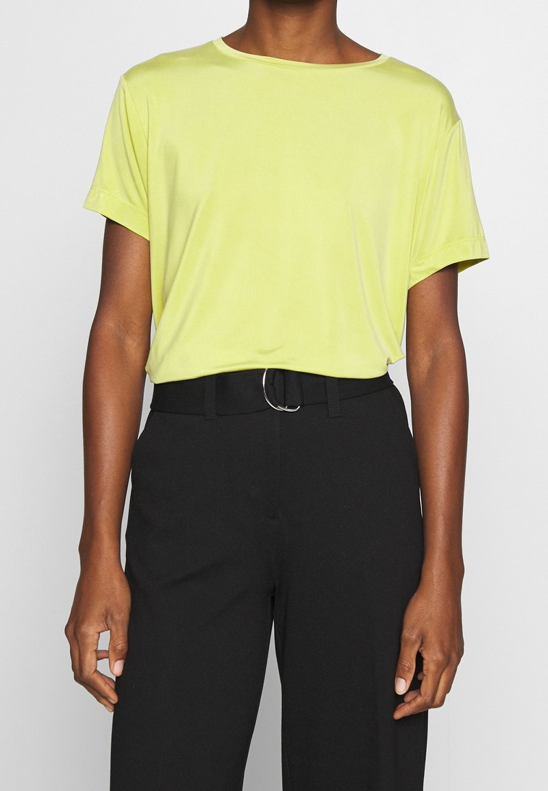 Personne portant une chemise jaune clair à manches courtes rentrée dans un pantalon noir taille haute avec une ceinture noire et une boucle argentée.
