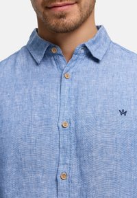 Man draagt lichtblauw linnen overhemd met knopen in beige en een klein donker geborduurd logo op de borst.