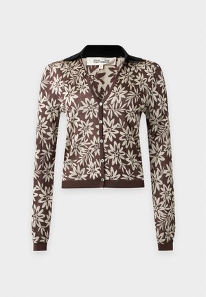 Femme debout, main sur la hanche, portant un cardigan à motifs floraux marron et beige et une jupe assortie fendue, ainsi que des sandales à talons noires.