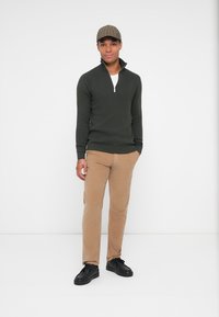 Tmavo zelený sveter na zips, béžové nohavice a čierne tenisky. Model má na hlave hnedú károvanú čiapku. Minimalistické pozadie.