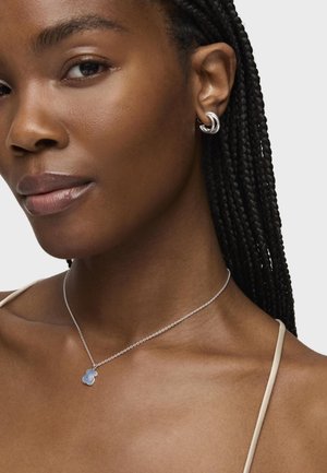 Collar de cadena de plata con un colgante azul, combinado con un aro de plata. Vista cercana del tono de piel y del cabello trenzado.