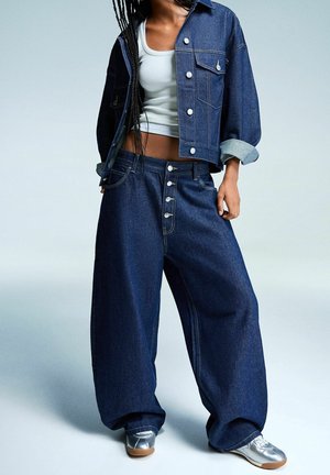 Persona vistiendo jeans sueltos de mezclilla azul oscuro, chaqueta corta a juego, top corto blanco y zapatillas plateadas sobre un fondo liso.