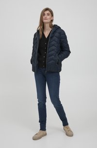 Doudoune marine avec capuche, fermeture éclair et motif en chevrons, associée à un cardigan noir, un jean bleu et des baskets beiges.