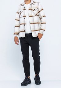 Randig flanellskjorta i vitt, brunt och beige, med två bröstfickor och svarta knappar, kombinerad med svarta avsmalnande byxor och svarta sneakers.