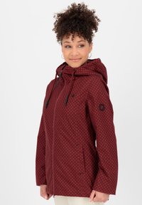 Rode jas met stippen, voorzien van een capuchon, ritsluiting en trekkoorden. Heeft twee zijzakken en zwarte hardware accenten.
