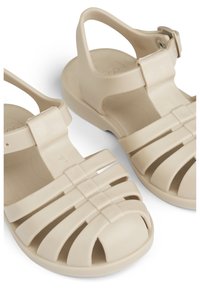 Beiget kumisaappaat, joissa on remmimäinen muotoilu ja avoin kärki. Kuvioitu pohja ja säädettävä nilkkaremmi, jossa sileä viimeistely.