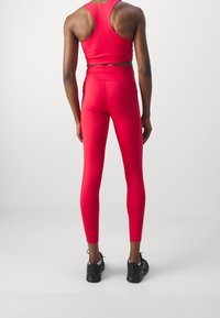 Czerwony sportowy biustonosz i legginsy z elastycznego materiału. Projekt ma dopasowany krój. Czarne buty sportowe dopełniają strój.