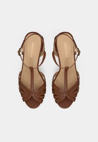 Sandales en cuir marron avec un design à fines lanières, comprenant des découpes courbes et une fermeture à boucle. Semelle intérieure beige douce avec le nom de la marque embossé.