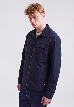 Junger Mann mit lockigen roten Haaren, der eine marineblaue Jacke mit Reißverschluss über einem karierten Hemd vor einem weißen Hintergrund trägt.