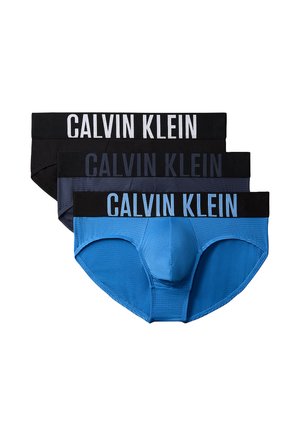 Wysoki, muskularny mężczyzna stoi boso na szarym tle, ubrany w ciemne slipy Calvin Klein, jedna ręka za plecami, neutralny wyraz twarzy.