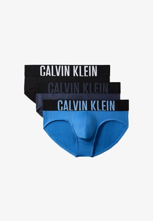 Lange, gespierde man staat op blote voeten tegen een grijze achtergrond, draagt donkere Calvin Klein slips, één hand achter de rug, neutrale uitdrukking.