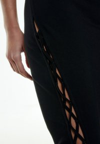 Zwarte jurk met een zijsplit, intrigerende vetersluiting en een soepele, rekbare stof. Close-up van het detail van de vetersluiting en de hand.