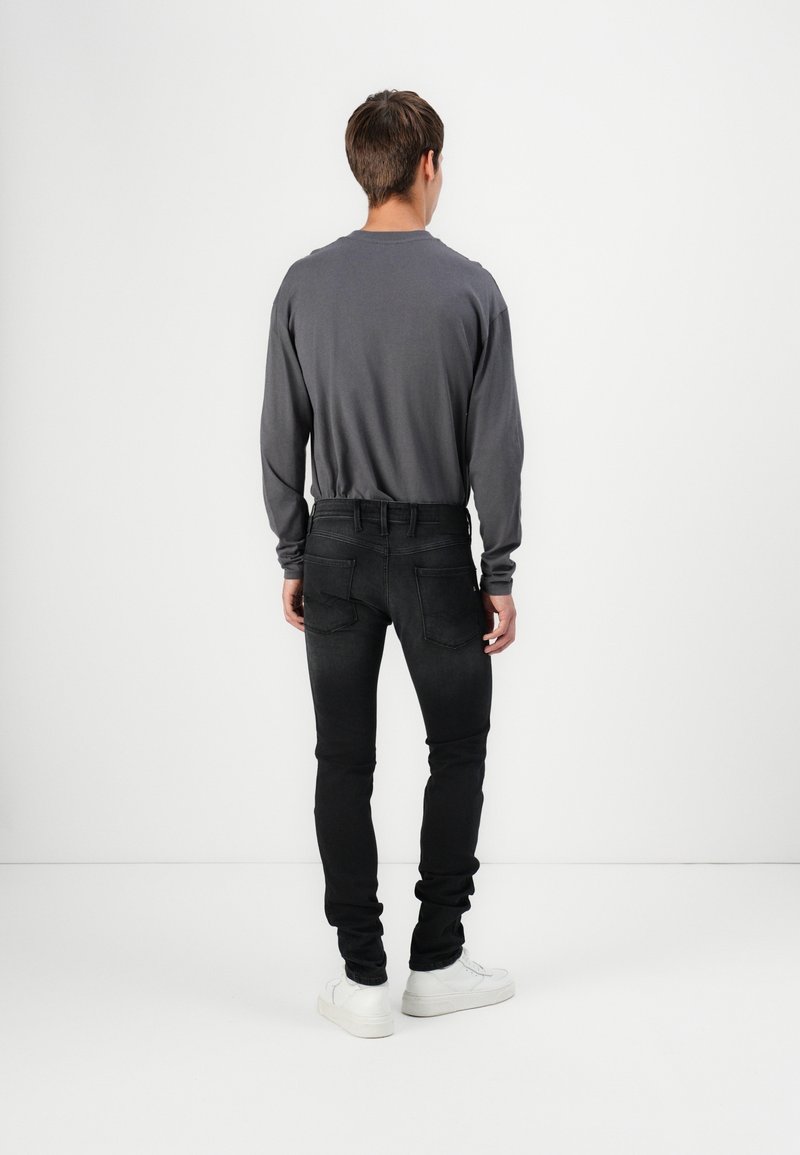 Homme debout, de dos, portant une chemise manches longues gris foncé, un jean slim noir et des baskets blanches sur fond blanc.