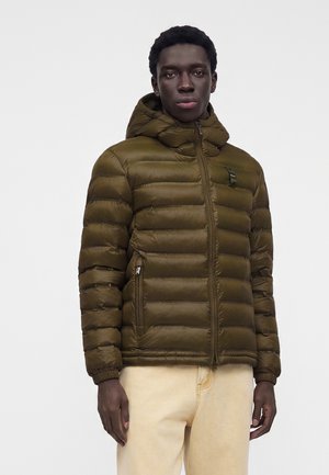 Jeune homme portant une doudoune à capuche marron et un pantalon beige, debout devant un fond clair uni.