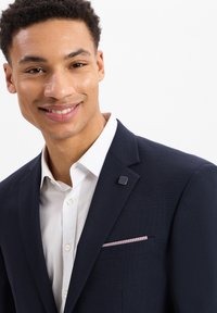 Granatowy blazer z teksturowanej tkaniny i klapami o wycięciu, z kontrastową białą koszulą pod spodem oraz dekoracyjnym wykończeniem kieszeni. Czarne akcenty metalowe.